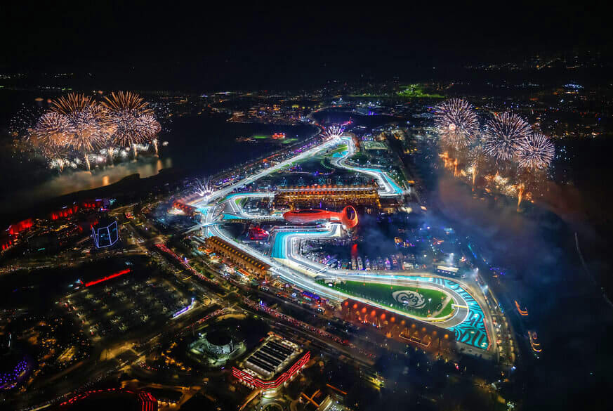Aerial night view of illuminated Formula 1 race track with blue lighting, fireworks, and city lights surrounding the circuit