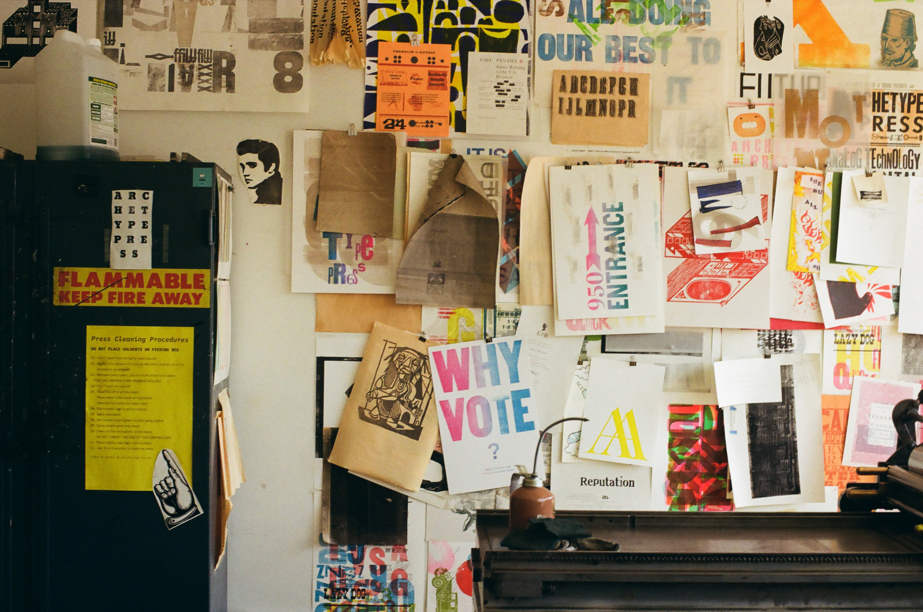 a collection of colorful letterpress prints pinned onto the wall. In the foreground is a letterpress machine