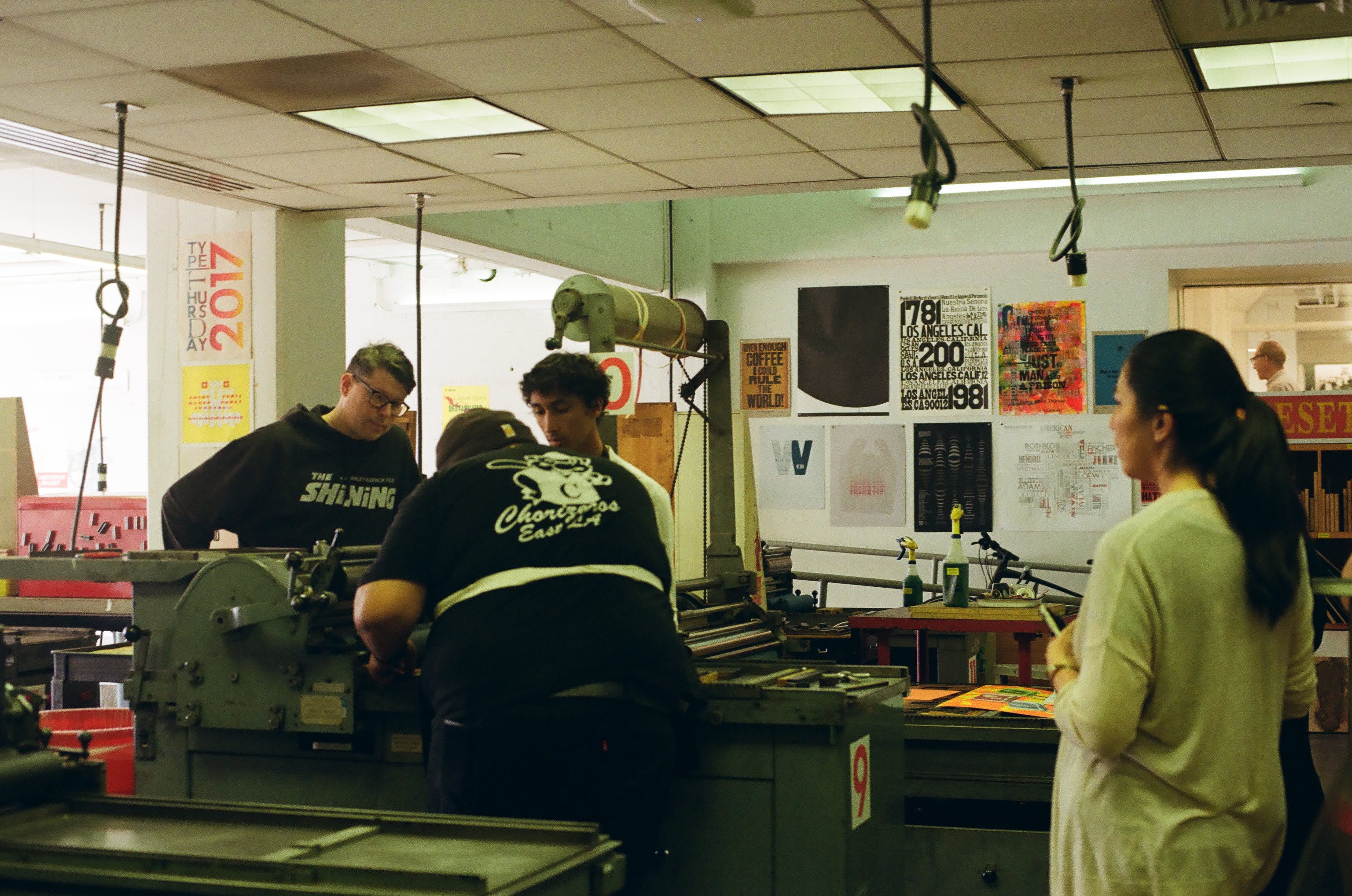 A few people gathered around a letterpress machine as it's being prepared.