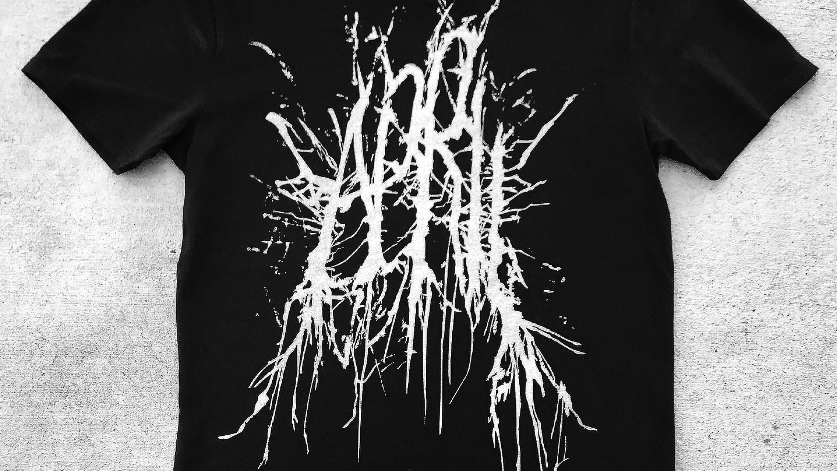 Top-down and close up view of the metal-inspired lettering on a tee shirt. The tee shirt is black, and the lettering is in white ink which has been screenprinted on.
