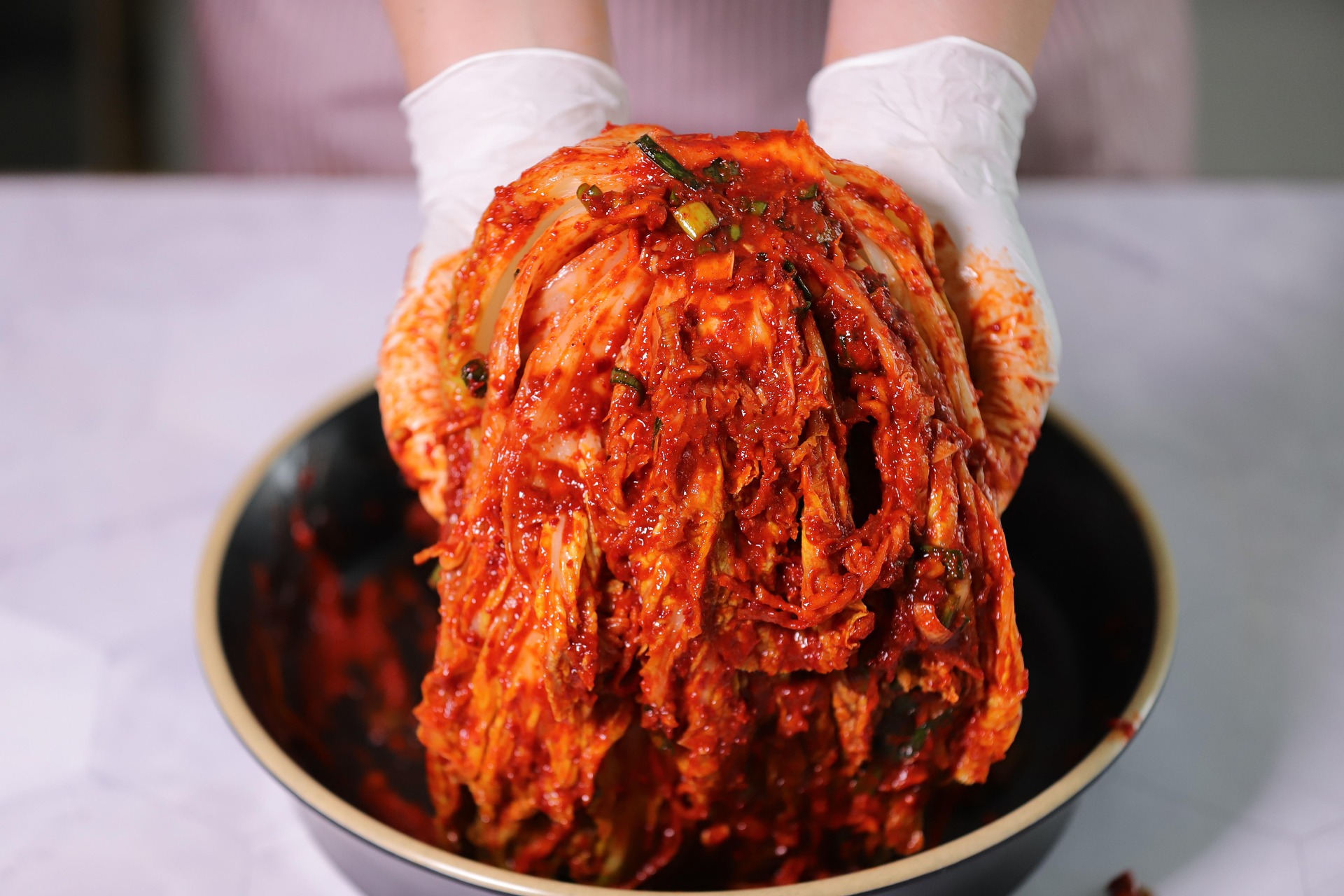유산균 덩어리 김치, 우리 부모님 건강 수명 늘리는 비법 대공개! 장갑 낀 손이 양념에 버무린 배추김치를 통째로 들어 올리는 모습 — 유산균 풍부한 발효 김치로 부모님 장건강과 식탁 건강을 챙기는 이미지