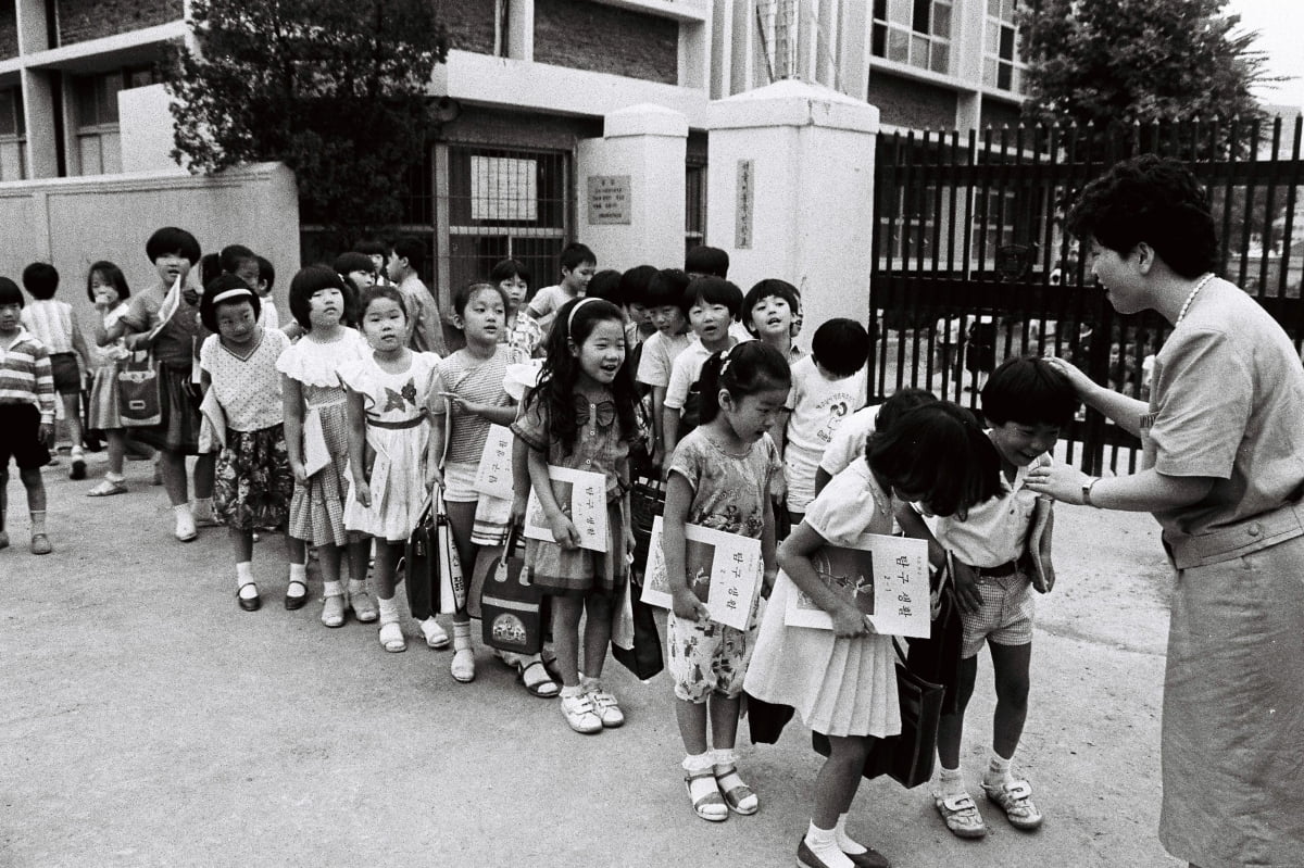 1970년대 국민학교 교문 앞에서 교사가 아이들을 줄 세우며 등교지도를 하는 흑백 사진—시니어 추억 회상 레트로 이미지