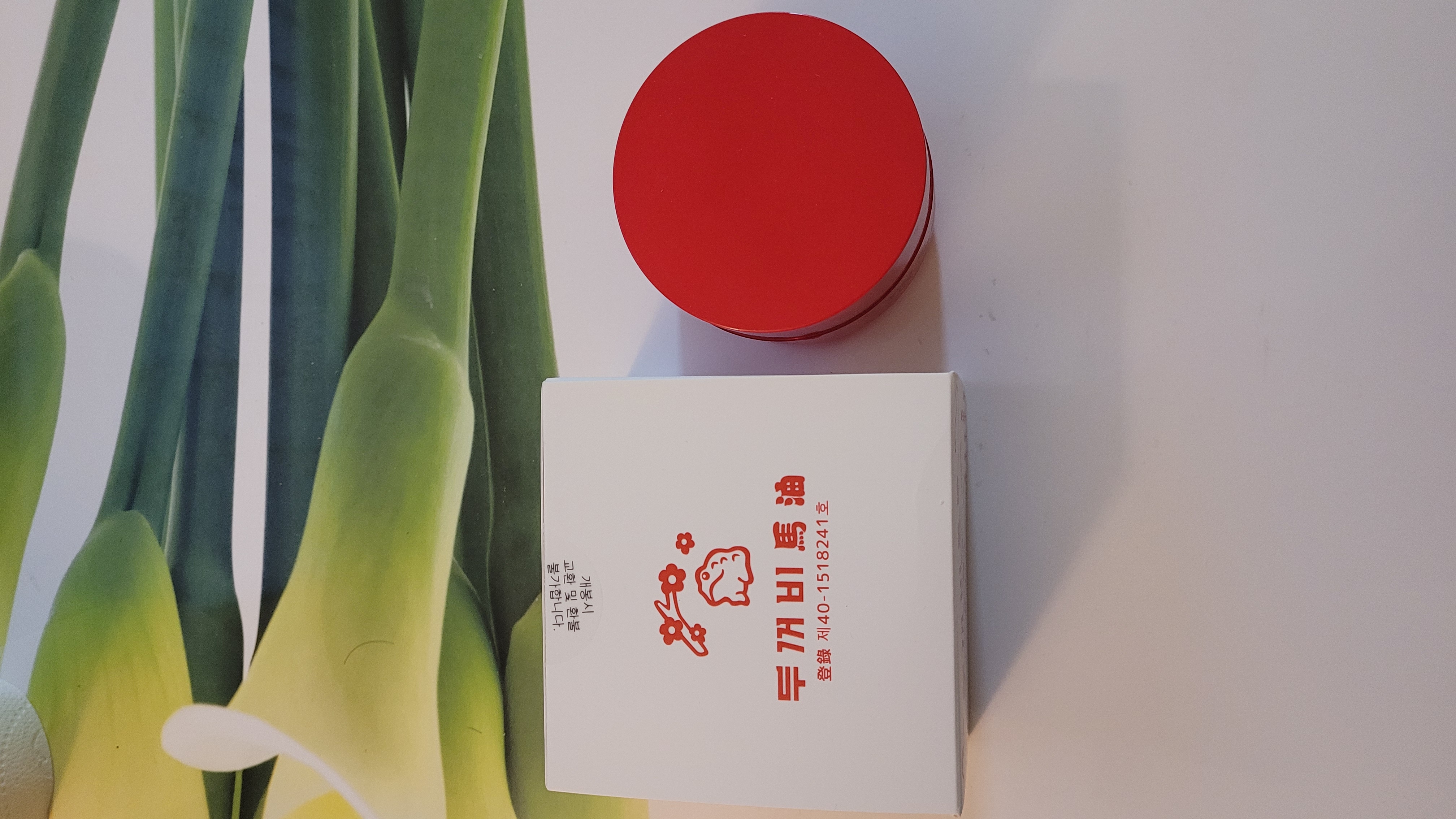 중국 전통 마유 크림 제품 이미지 - 흰색 박스에 붉은색 글씨로 '두개비마유' 한자가 적혀있고 빨간 원형 용기가 함께 놓여있으며 배경에 자연스러운 녹색 식물 잎이 보이는 천연 화장품 컨셉 사진