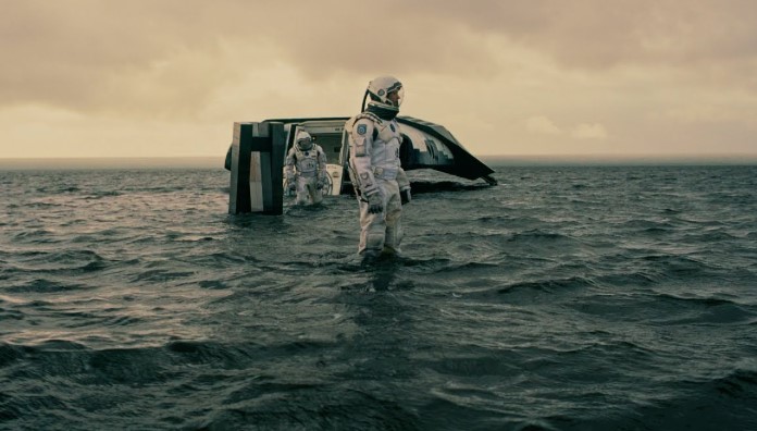 인터스텔라 밀러 행성 장면, 쿠퍼와 브랜드 박사가 우주복을 입고 거친 파도가 치는 외계 바다를 걸어 나오고 있으며 뒤편에는 착륙선이 반쯤 잠겨 있습니다