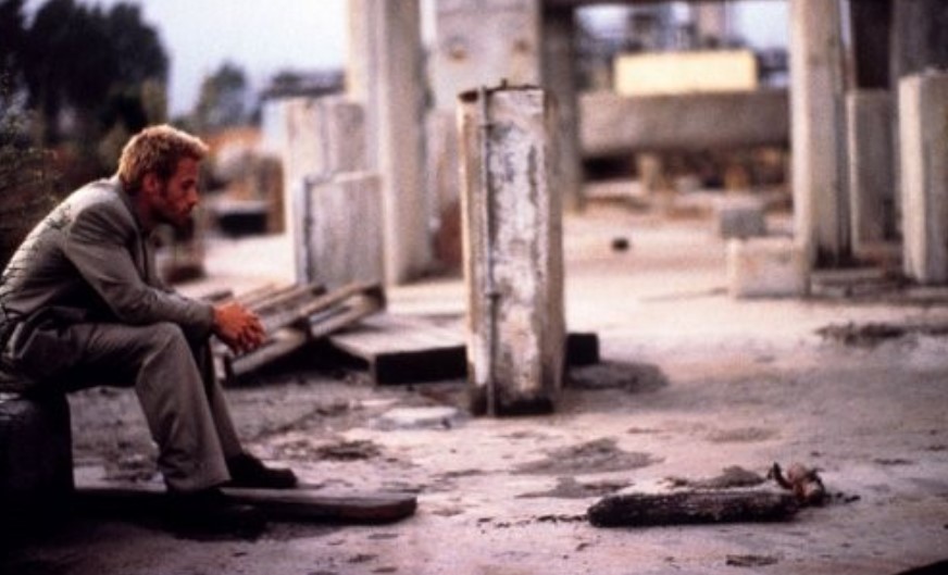 메멘토 영화 스틸컷 - 폐허가 된 건물 잔해 속에 레너드(가이 피어스)가 홀로 앉아 허공을 바라보는 장면, 기억도 진실도 없는 고독한 비극을 상징