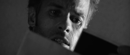 메멘토 영화 흑백 스틸컷 - 어둠 속에서 극도로 불안하고 혼란스러운 표정으로 무언가를 응시하는 레너드(가이 피어스) 클로즈업, 망상과 진실의 경계를 상징