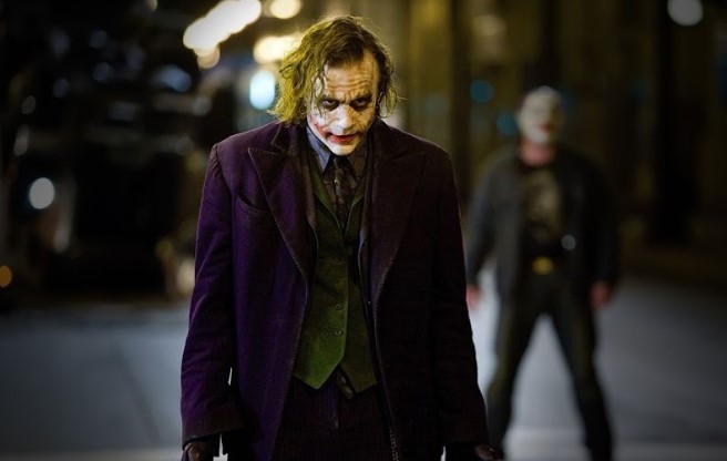 다크 나이트 야간 거리 장면 - 히스 레저의 조커가 고개를 숙인 채 어두운 거리를 걸어가고, 뒤편으로 배트맨이 흐릿하게 서 있는 모습. 조커와 배트맨이 서로를 완성시키는 동전의 양면 관계를 상징적으로 보여주는 장면