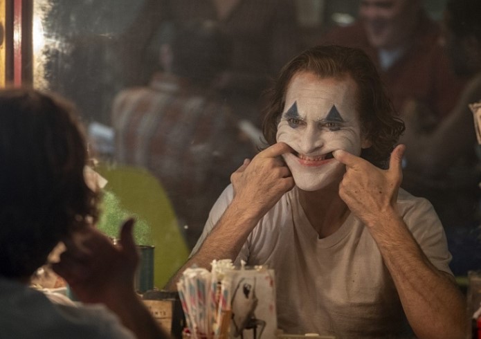 영화 조커(2019) 호아킨 피닉스가 흰 광대 분장을 한 채 거울 앞에서 두 손가락으로 입꼬리를 억지로 올려 웃는 표정을 만드는 장면으로 아서 플렉의 무너진 정체성과 내면의 고통을 상징하는 소름 돋는 명장면