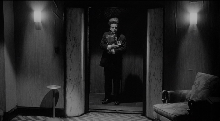 이레이저헤드(1977) 주인공 헨리 스펜서가 어두운 복도 문 앞에서 서 있는 스틸컷 — 데이비드 린치의 필라델피아 시절이 반영된 황폐한 산업 도시 풍경