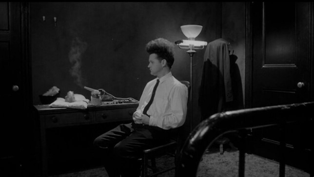 이레이저헤드(1977) 주인공 헨리 스펜서가 어두운 방 안 의자에 홀로 앉아 기형 아기를 바라보는 스틸컷 — 고립과 부성 공포를 상징하는 장면