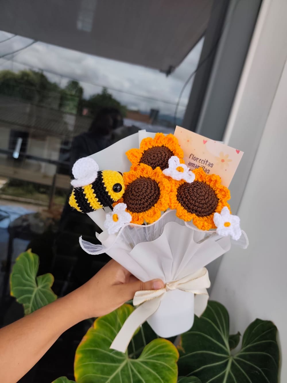 Ramo compuesto por tres girasoles grandes tejidos a crochet, adornado con una abejita tejida a rayas amarillas y negras, tres margaritas blancas pequeñas, envuelto en papel floral blanco con un moño grueso color crema.