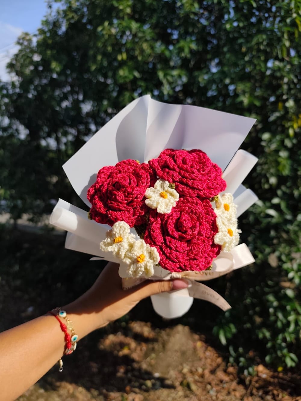 Fotografía en primer plano de un ramo tejido a crochet compuesto por tres rosas rojas vibrantes y cinco pequeñas margaritas blancas con centro amarillo. Las flores están envueltas en papel floristero blanco mate y sujetadas por una cinta de tela color crema con la palabra 'Love' impresa. Fondo de naturaleza desenfocado.