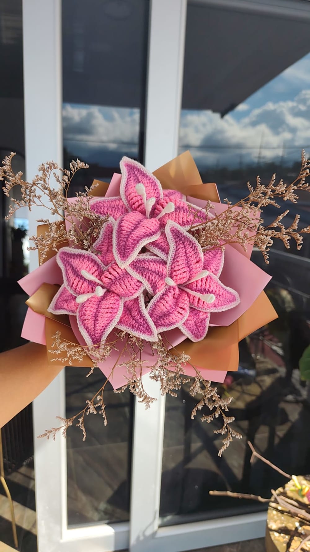 Elegante bouquet artesanal compuesto por tres lirios tejidos a crochet en color rosa vibrante con bordes blancos definidos y estambres realistas. Incluye ramas de flores secas decorativas para un toque rústico. Envuelto en capas de papel rosa mate y dorado metalizado.