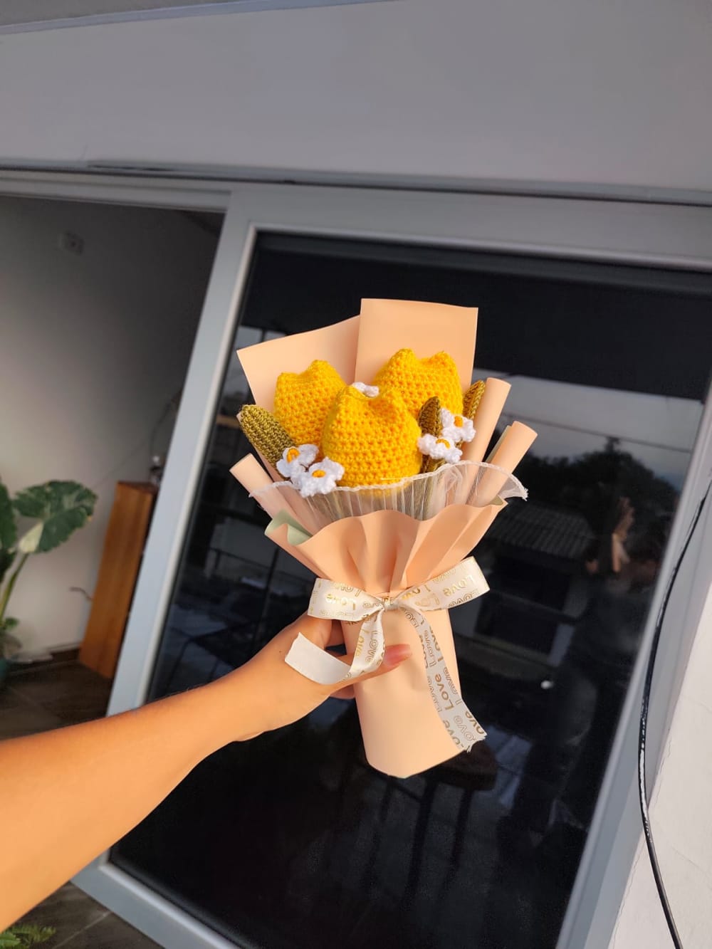 Ramo decorativo tejido artesanalmente a mano que incluye tres tulipanes amarillos vibrantes acompañados de pequeñas margaritas blancas con centro amarillo. El conjunto está envuelto en papel decorativo color melocotón suave y atado con un elegante moño de cinta color crema con la inscripción 'Love' dorada.