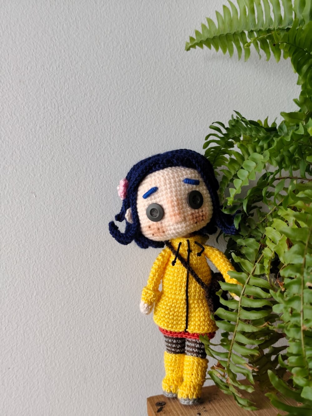 Detallada muñeca amigurumi de Coraline Jones tejida a mano a crochet. Luce su característico cabello azul oscuro con una flor rosa, impermeable amarillo brillante, botas amarillas y leggings a rayas. Destacan sus icónicos ojos de botón negros. Posada junto a un helecho verde natural sobre un fondo blanco.
