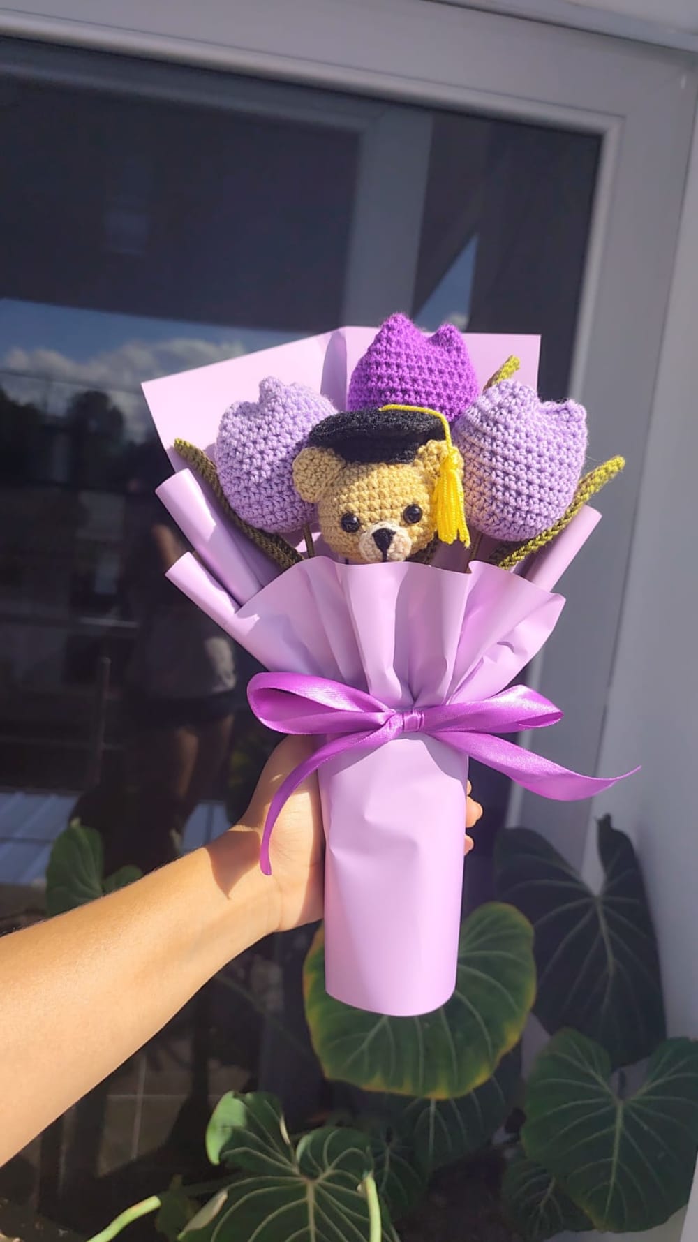 Ramo de graduación artesanal tejido a mano a crochet que presenta tres tulipanes púrpuras y un tierno oso amigurumi con su birrete negro de graduado y borla amarilla. Todo el conjunto está envuelto en papel morado suave y decorado con un impecable moño de cinta morada satinada.
