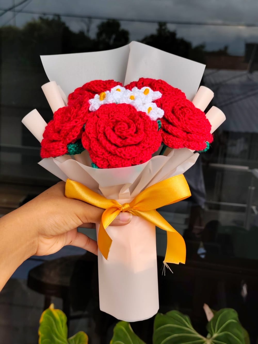 Fotografía de un ramo artesanal tejido a mano a crochet con rosas rojas vibrantes y pequeñas margaritas blancas. El ramo se encuentra envuelto en un pulcro papel blanco mate y destaca por un gran moño de cinta satinada color amarillo oro brillante. Capturado en un ambiente interior iluminado.