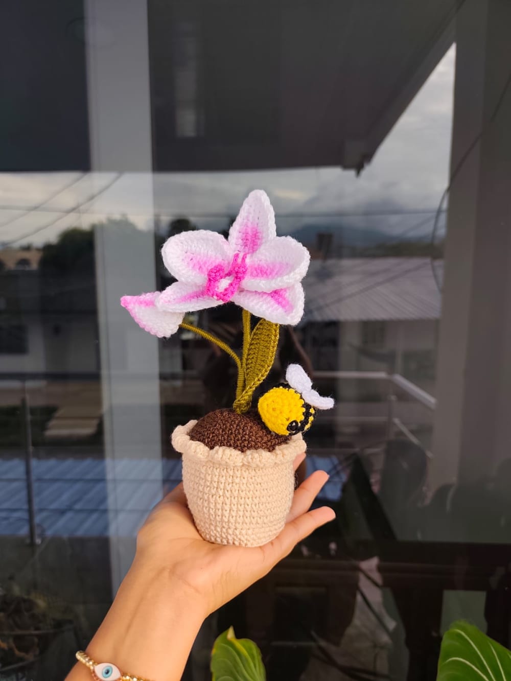 Fotografía de una mini maceta artesanal tejida a crochet en color beige, que sostiene una elegante orquídea blanca con centros degradados en rosa fucsia. Una pequeña y tierna abejita amigurumi está posada sobre la base de la maceta. Un detalle botánico ideal para decoración de interiores y escritorios.