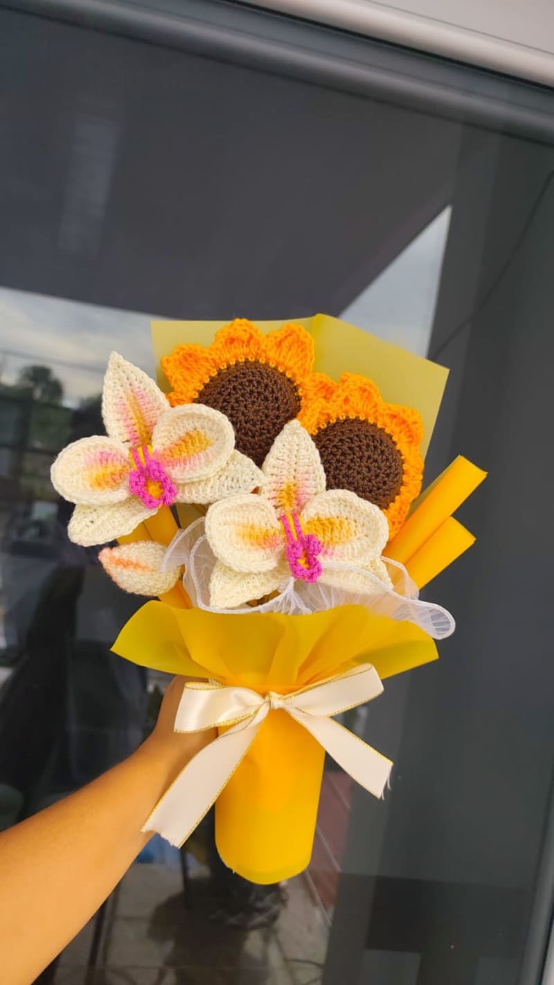 Ramo tejido a crochet que combina dos girasoles y dos orquídeas blancas con centro fucsia, envuelto en papel amarillo floral y atado con un moño satinado crema.