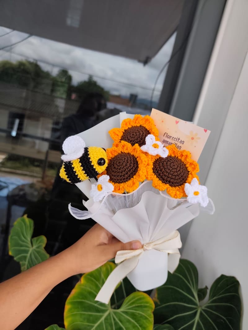 Ramo compuesto por tres girasoles grandes tejidos a crochet, adornado con una abejita tejida a rayas amarillas y negras, tres margaritas blancas pequeñas, envuelto en papel floral blanco con un moño grueso color crema.