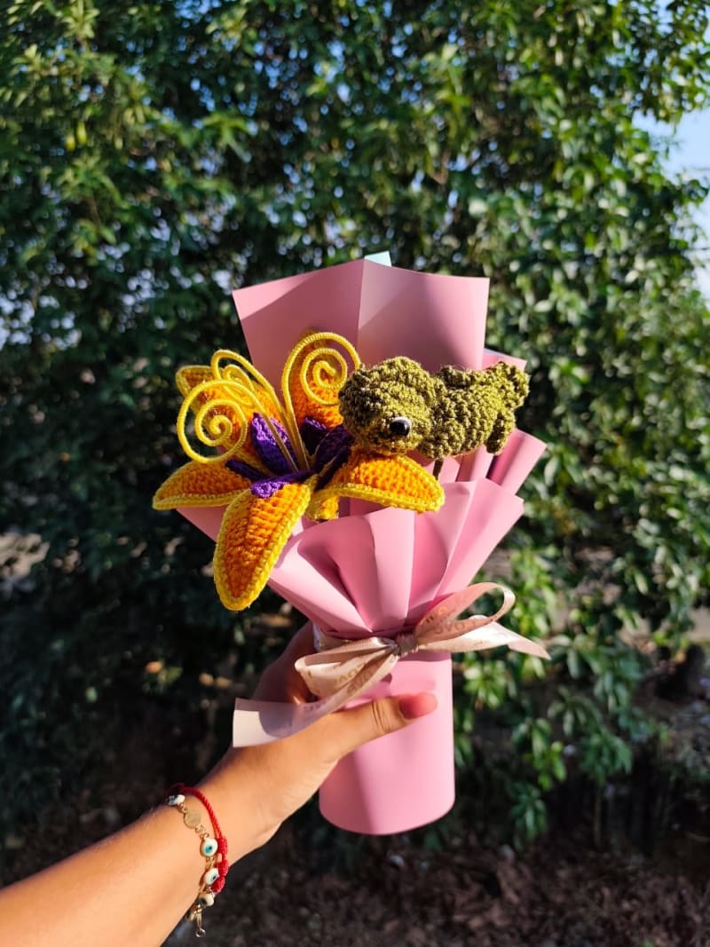 Primer plano de un ramo tejido a mano que muestra un gran lirio amarillo con detalles púrpuras y un pequeño camaleón verde amigurumi tejido con gran detalle, posado sobre la flor. Envuelto en papel rosado con un moño satinado crema, bajo luz natural de sol.