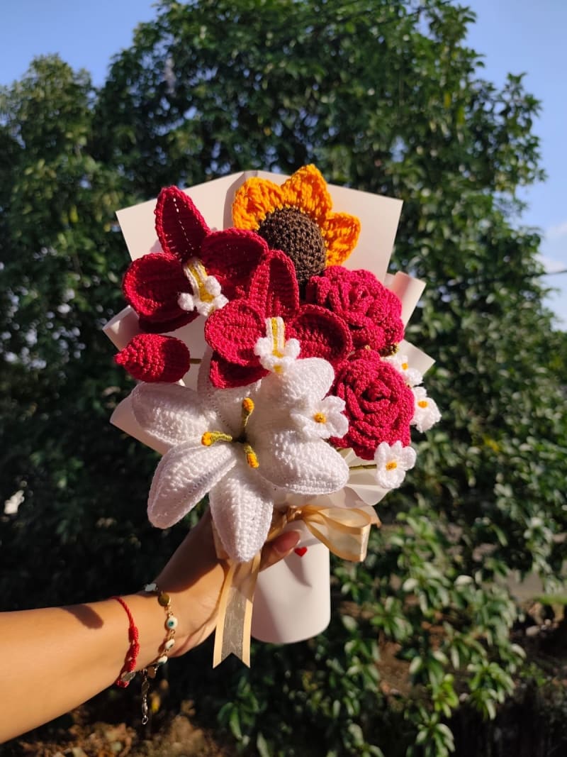 Fotografía en primer plano de un lujoso ramo tejido a crochet que incluye orquídeas borgoña, un gran lirio blanco con estambres detallados, rosas a juego y un vibrante girasol. Decorado con pequeñas margaritas blancas, envuelto en papel crema satinado y finalizado con un moño dorado elegante. Luz natural exterior.