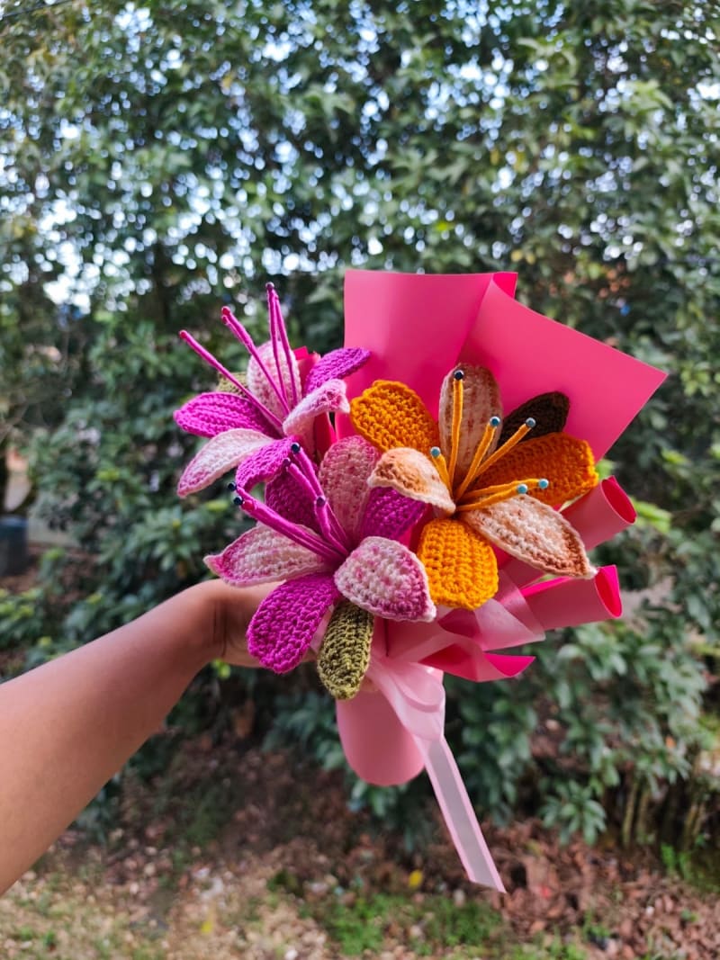 Vibrante ramo tejido a mano que presenta varios lirios de crochet detallados en tonos fucsia intenso y naranja melocotón con degradados. Las flores destacan por sus estambres largos con puntas de colores. Está envuelto en papel rosado fucsia mate y atado con una cinta rosada clara satinada