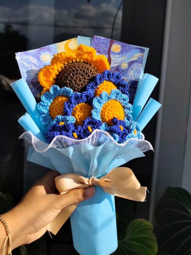 Fotografía de un sofisticado ramo artesanal tejido a crochet inspirado en la obra de Vincent van Gogh. Destaca un gran girasol central acompañado de flores azules y amarillas que simulan los giros de "La Noche Estrellada". El ramo incluye una tarjeta de la pintura original de fondo y está envuelto en papel azul cielo con un elegante moño satinado champagne.