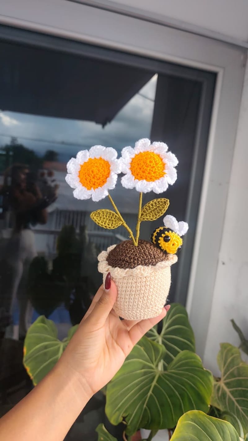 Macetita decorativa tejida a mano color beige y tierra marrón, de la cual brotan dos tulipanes amarillos a crochet con hojas verdes. En el borde de la maceta hay una pequeña abejita amigurumi posada asomándose.