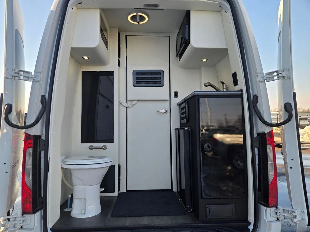 Sprinter rear bathroom viewed through open rear doors, with a visible toilet and cabinetry