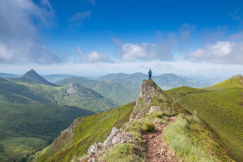 Randonnée monts du Cantal : homme de dos sur le sommet d'une montagne verdoyante, entouré de monts