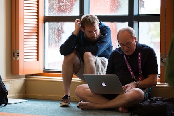 One sitting on the ground with their laptop and another person leaning over to look at their screen.