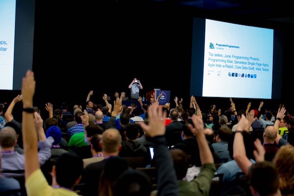 A conference hall where many are raising their hands and the speaker taking a picture of the audience