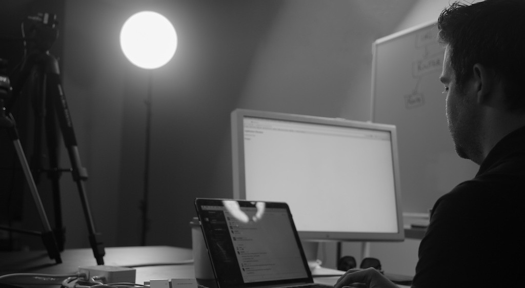 A person with a laptop and a monitor sitting by a whiteboard and desk