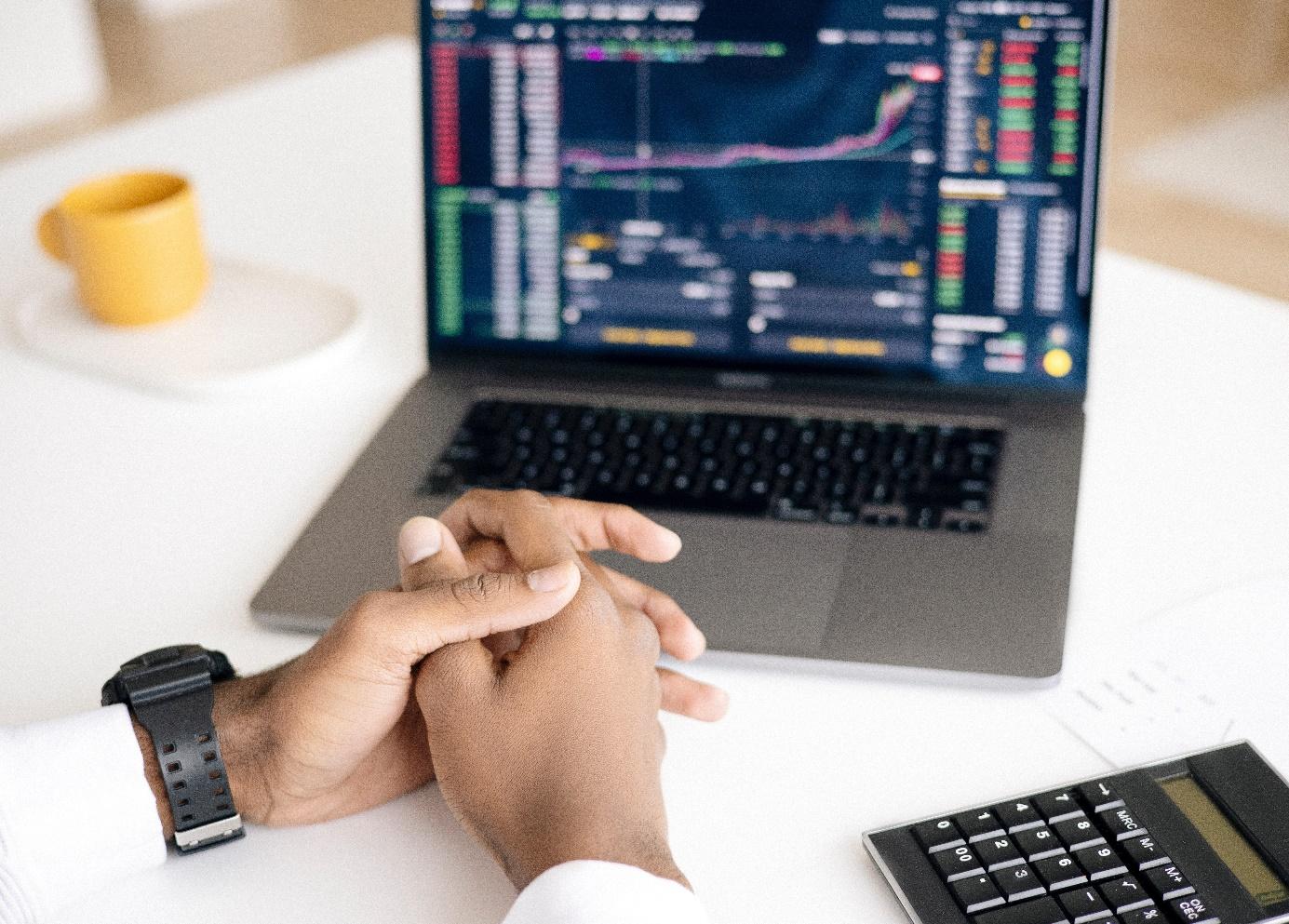 View of an opened laptop on a white desk next to a yellow coffee cup and a calculator. A male person sits in front of it with folded hands. The question of when to use fixed or dynamic pricing is on the minds of many retailers. You can get answers to this question in this article!