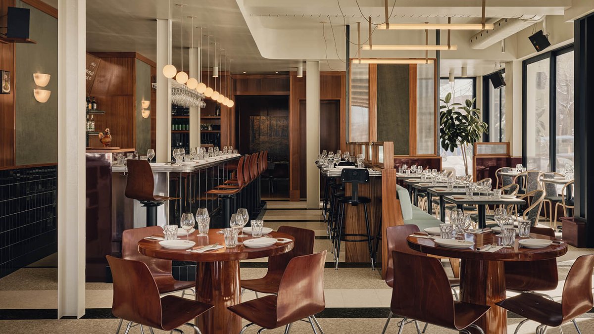 Restaurant interior in Montreal featuring wooden tables, curved chairs, and a bar with stools under globe pendant lights.