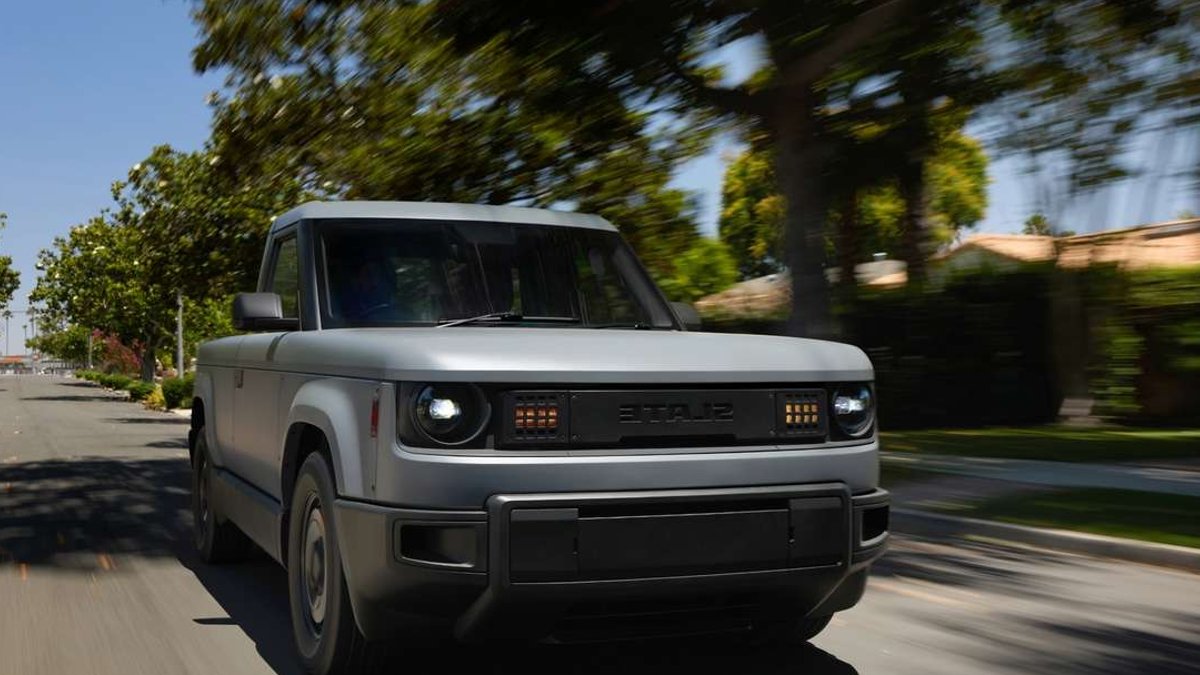 Gray Slate electric pickup truck driving along a tree-lined residential street, showing its front grille, LED headlights, and