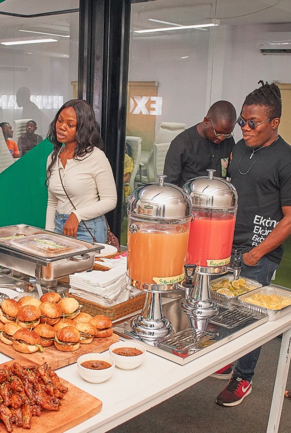 a group of people standing around a buffet table with sandwiches and juices on the side of the table