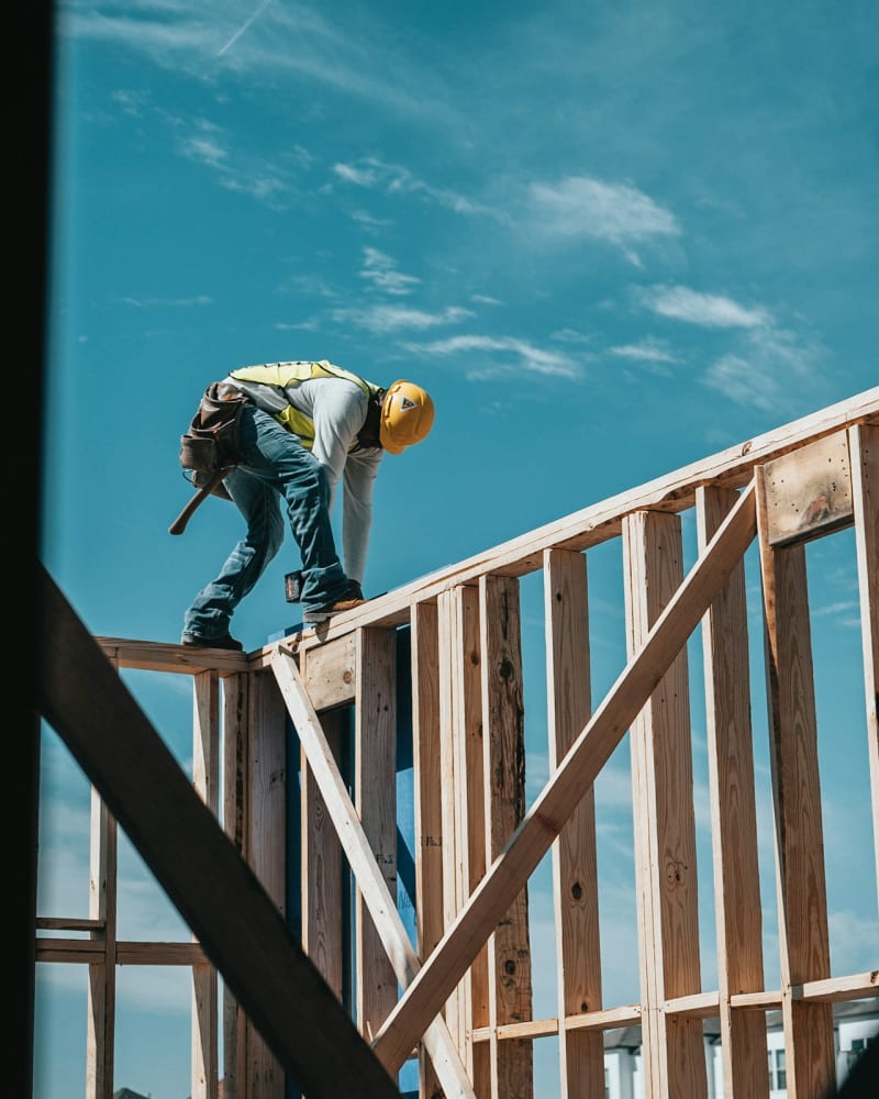 A builder in a hard hat and hi-vis working on a timber frame against a clear sky
