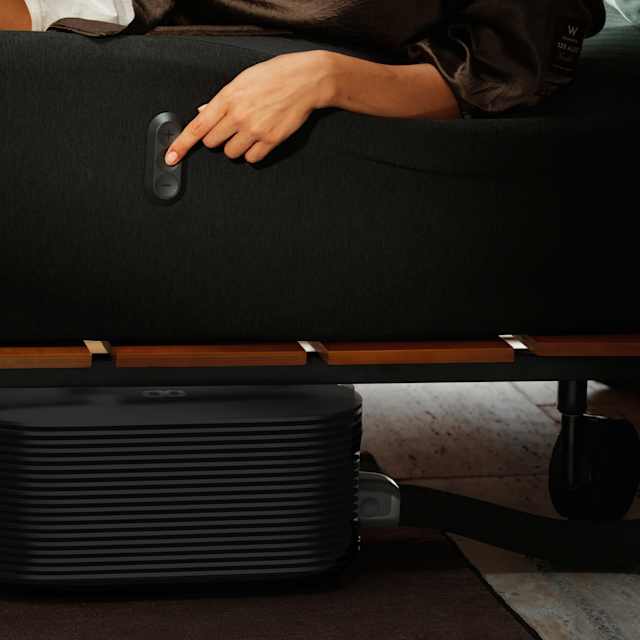 A hand pressing the temperature buttons on the side of the Pod 6 Cover, with the Hub under the bed frame.