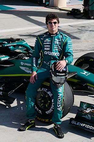 Lance Stroll in team kit kneeling beside the Aston Martin car in the pit lane