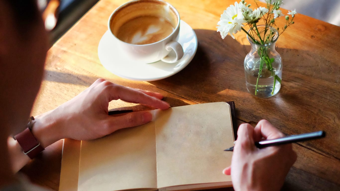Kreativer werden mir den drei Morgenseiten Eine Person schreibt in ein leeres Tagebuch auf einem Holztisch neben einer Tasse Kaffee und einer kleinen Blumenvase.