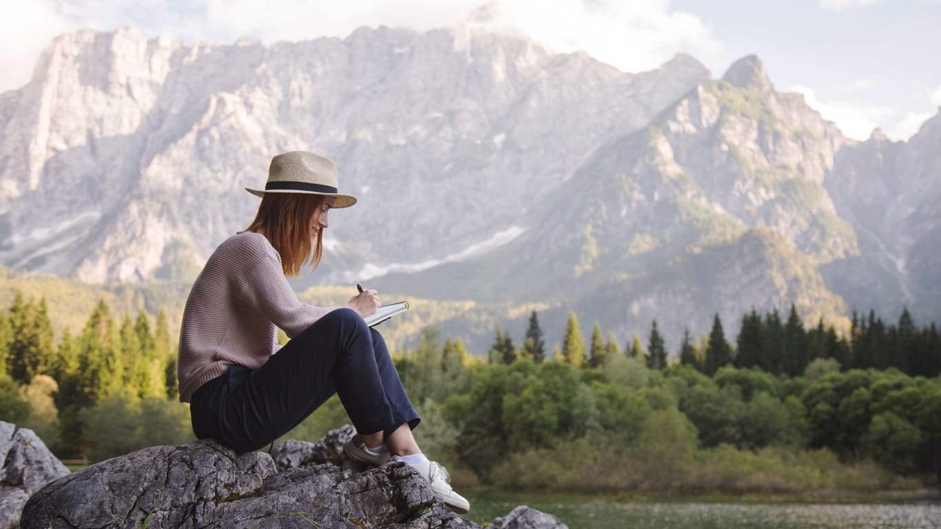 Auszeit im Alltag Eine Frau sitzt auf Felsen am Ufer eines Sees und schreibt in ein Notizbuch, im Hintergrund sind Berge und Bäume zu sehen.