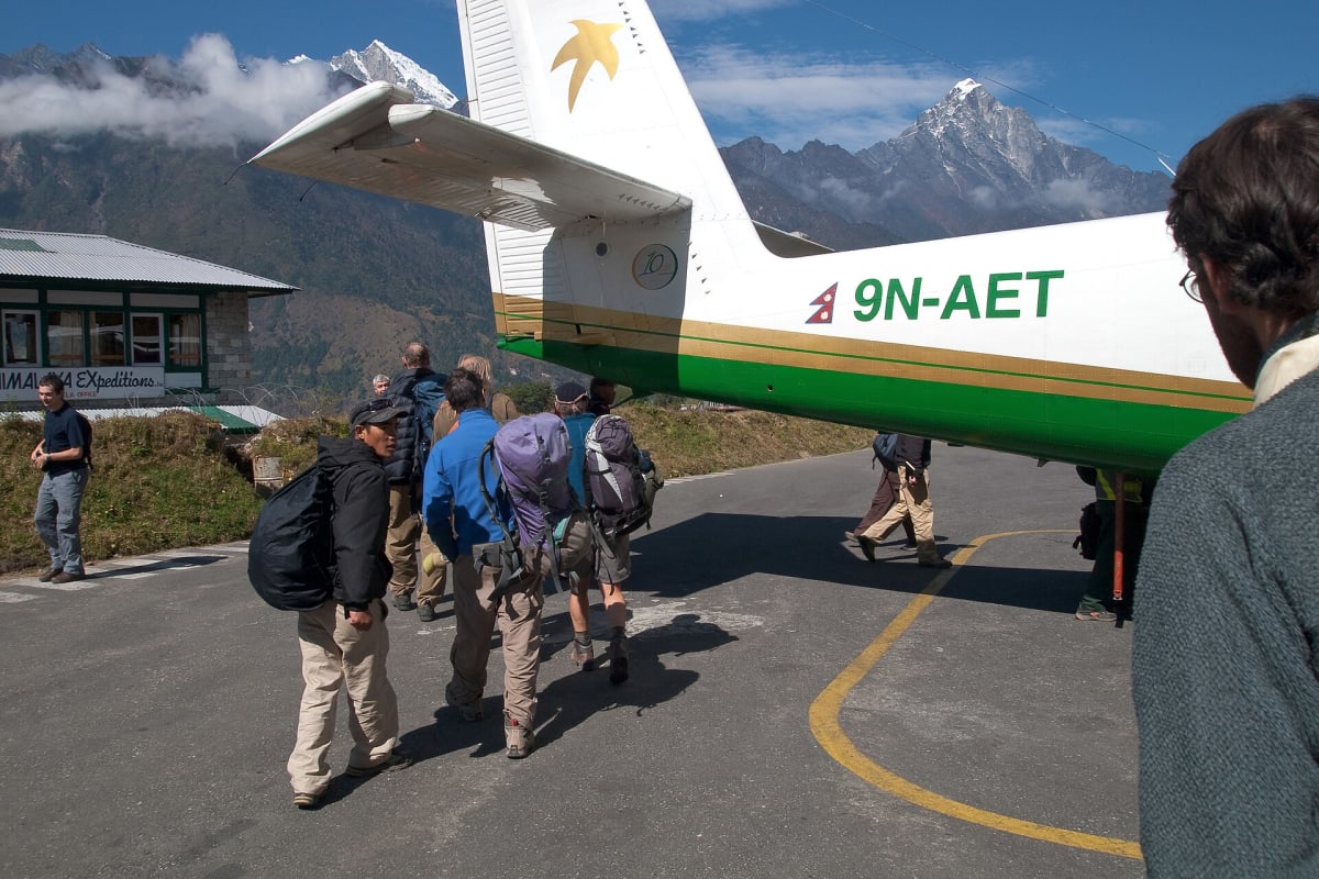 Trekkers with rucksacks walking across the apron towards a Twin Otter aircraft at Lukla airport, with a snow peak beyond.