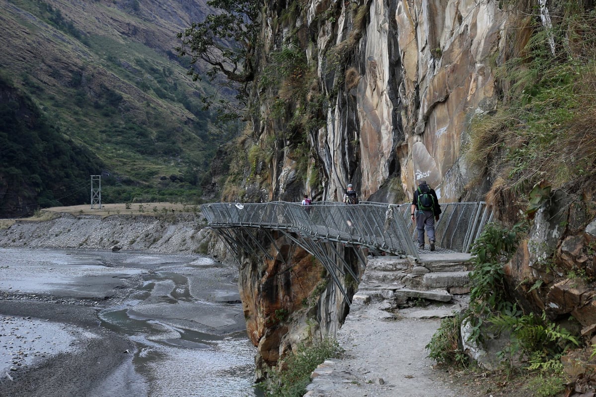 Trekkers on a steel walkway bolted to a cliff above the gravel bed of the Budhi Gandaki between Yaru Bagar and Jagat.