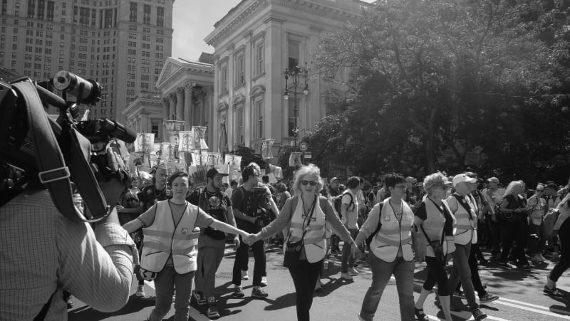 News cameras cover the front of the march as volunteers in vests clear the way