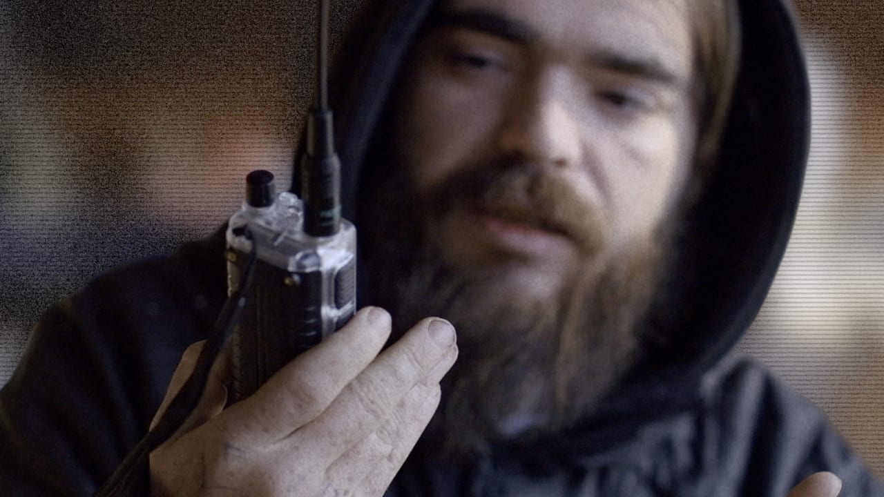 Man with long hair and beard holding a handheld radio transceiver, looking downward.