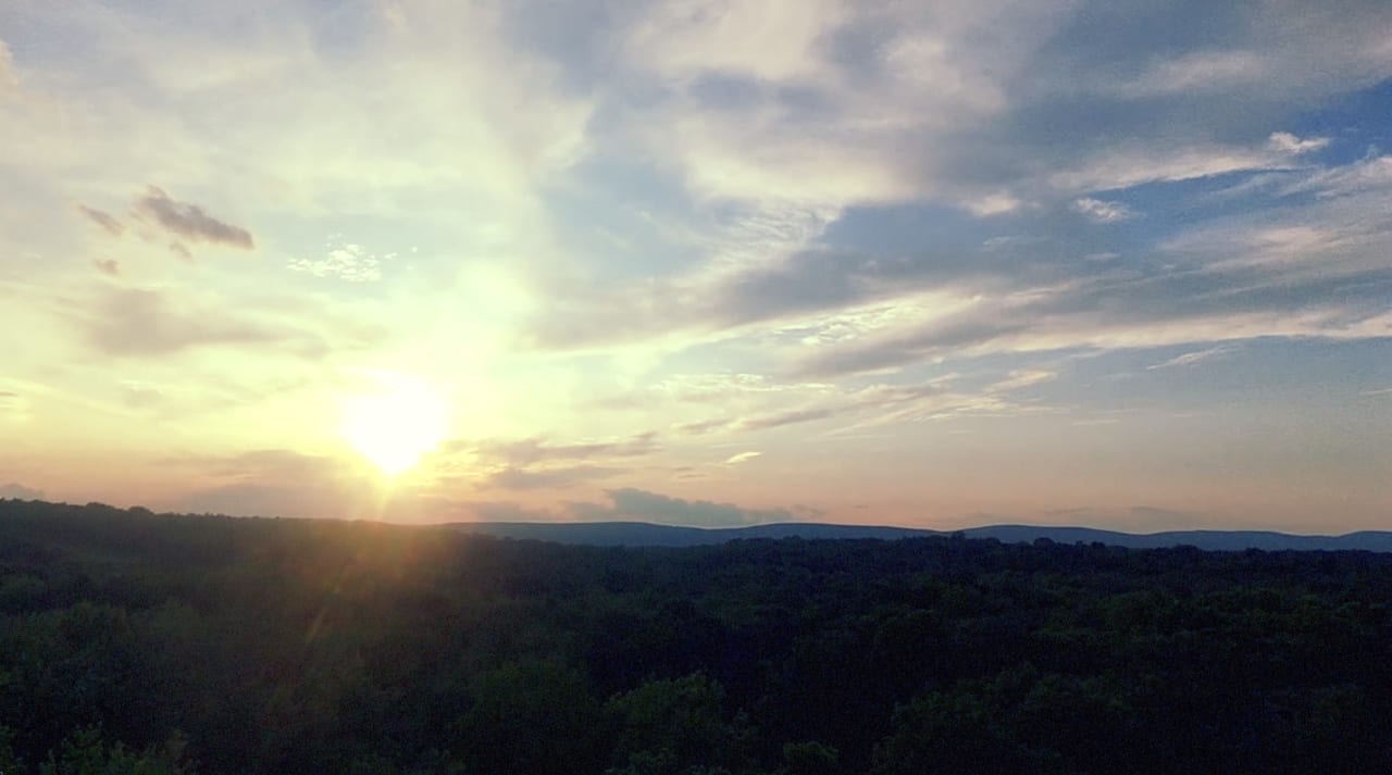 Sunset over a forested valley with distant mountains under partly cloudy sky.