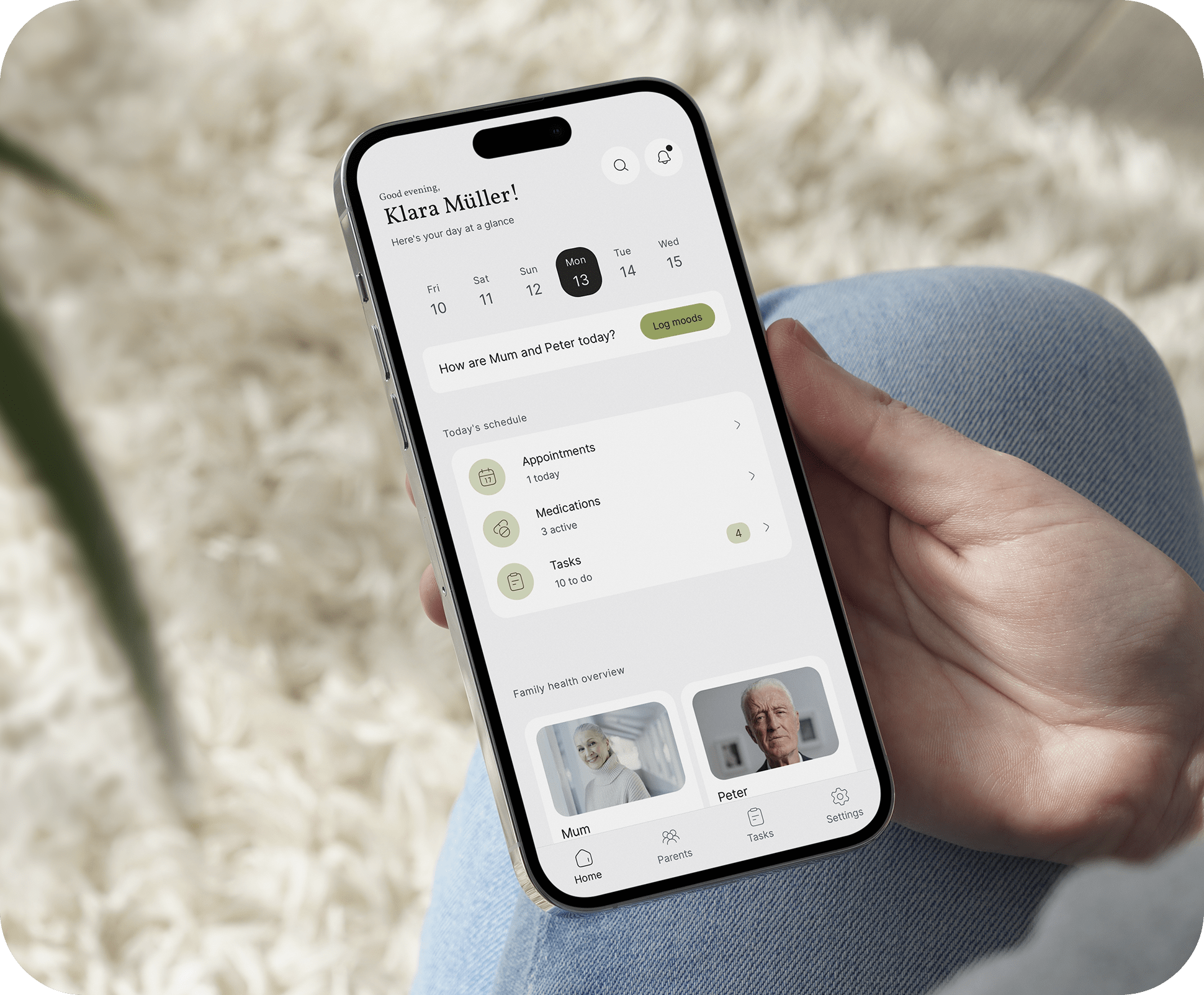 Hand holding an iPhone showing the Kinlog family caregiving app home dashboard with day at a glance, appointments medications and tasks summary, plus Mum and Peter care profiles