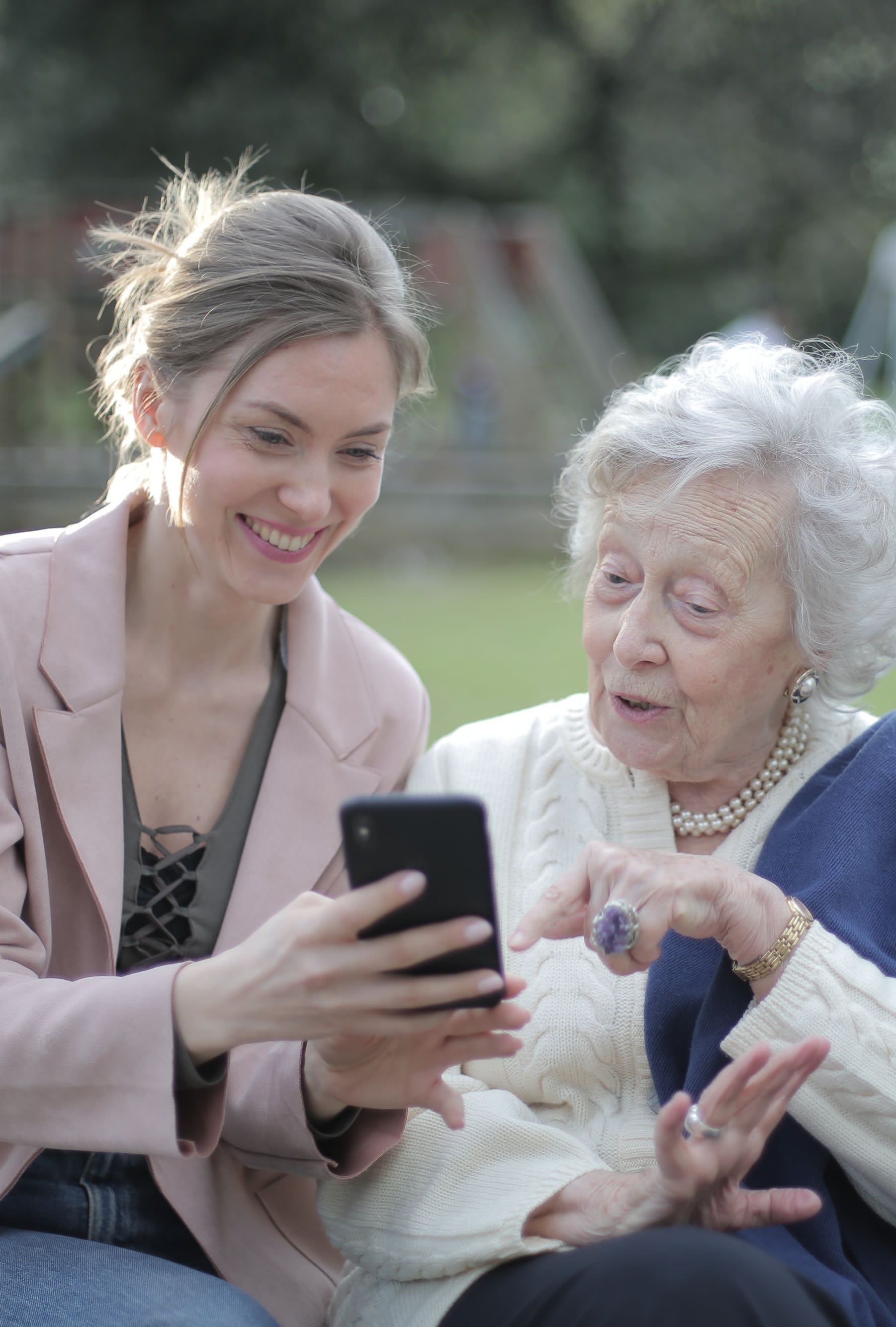 A daughter showing her mum something on her phone, both smiling, on a bench outside